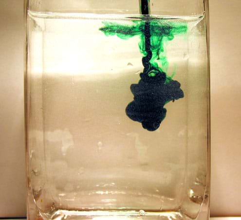 Photo shows plume of green food coloring dropping deep into water in a glass container.