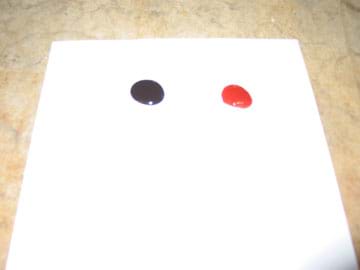 Photo shows a dark drop next to a red drop on a white piece of paper.