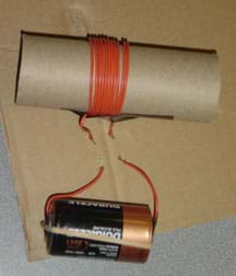 A photo shows a cardboard tube wrapped in wire and placed on a piece of flat cardboard. Two long tails of the coiled wire run through holes in the cardboard and are attached with a rubber band to opposite ends of a D-cell battery.